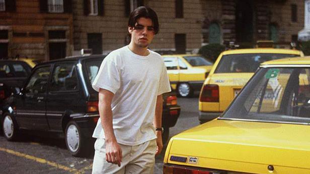 Ein junger Mann in weißem T-Shirt steht auf der Straße, im Hintergrund parkende Autos, darunter gelbe Taxis.