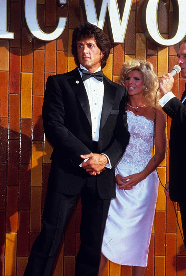 Sylvester Stallone und seine Begleitung posieren vor einer Wand mit dem Schriftzug „Hollywood“.