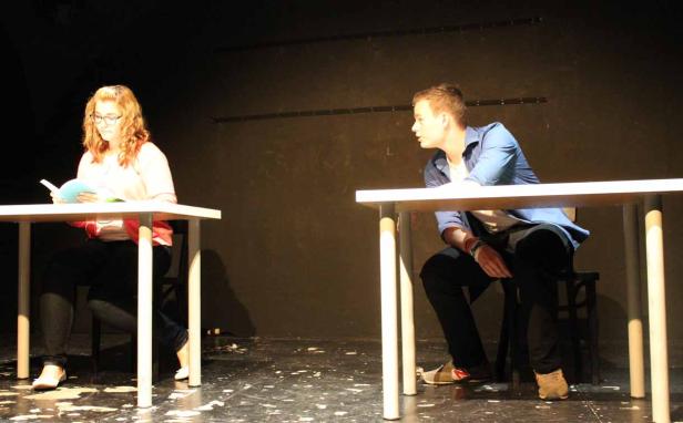 Eine junge Frau liest ein Buch, während ein junger Mann auf der Bühne sitzt und wegschaut.