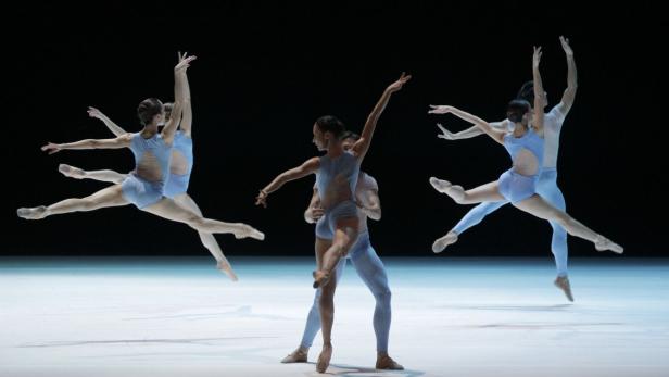 Mehrere Balletttänzer und -tänzerinnen in blauen Kostümen führen eine synchrone Choreografie auf.