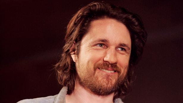 Ein lächelnder Mann mit braunen, gewellten Haaren und Bart vor einem dunklen Hintergrund.