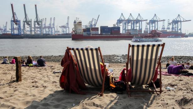 Menschen entspannen an einem Strand in Hamburg mit Blick auf Containerschiffe und Kräne.