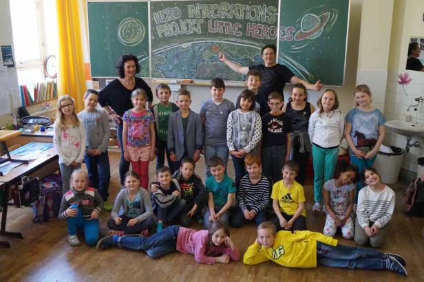 Eine Schulklasse posiert mit ihren Lehrern vor einer Tafel mit der Aufschrift „Hero Integrations Projekt Little Heroes“.