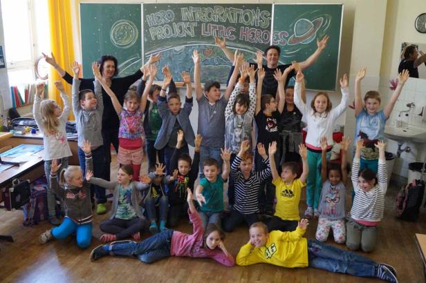 Eine Schulklasse und zwei Lehrer posieren mit erhobenen Händen vor einer Tafel.