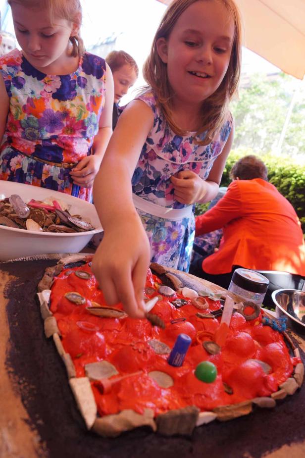 Ein Mädchen verziert eine rote, pizzaähnliche Oberfläche mit verschiedenen Objekten.