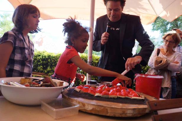 Ein Mädchen verziert unter Aufsicht einen großen Kuchen mit roten Elementen.