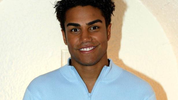 Ein lächelnder junger Mann mit dunklen Haaren und hellblauem Pullover.