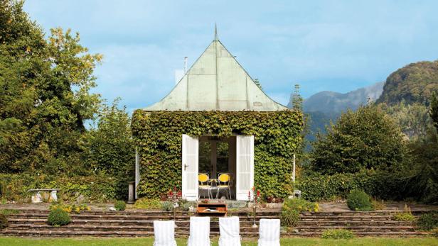 Ein kleines, mit Efeu bewachsenes Gartenhaus mit weißen Stühlen davor.