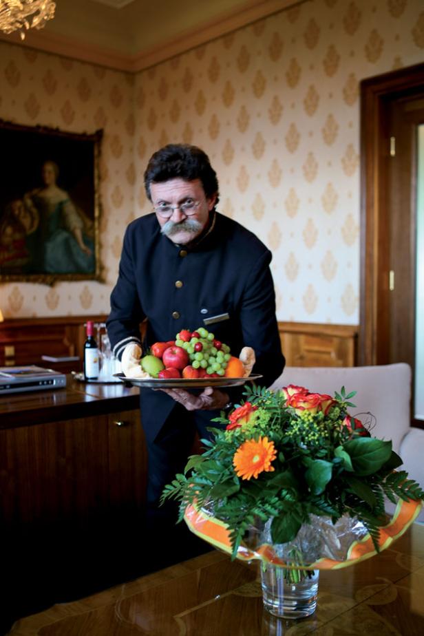 Ein Hotelangestellter mit Schnurrbart serviert eine Obstplatte in einem eleganten Zimmer.