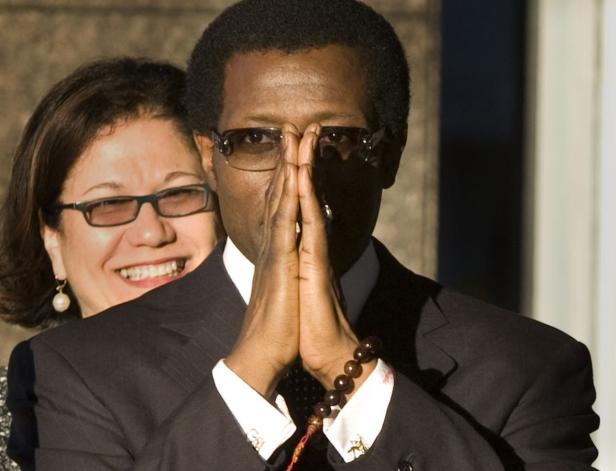 Ein Mann mit Brille hält die Hände vor dem Gesicht, während eine Frau im Hintergrund lächelt.