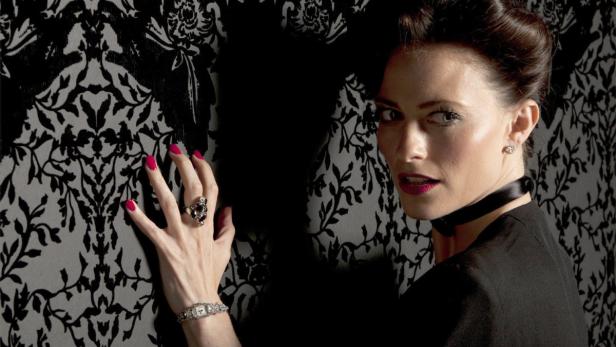 Eine Frau mit dunklem Haar, Schmuck und schwarzem Choker vor einer gemusterten Tapete.