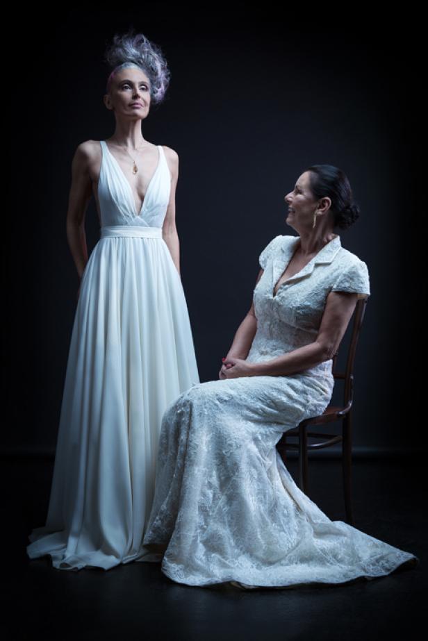 Zwei Frauen in weißen Kleidern posieren vor einem dunklen Hintergrund.