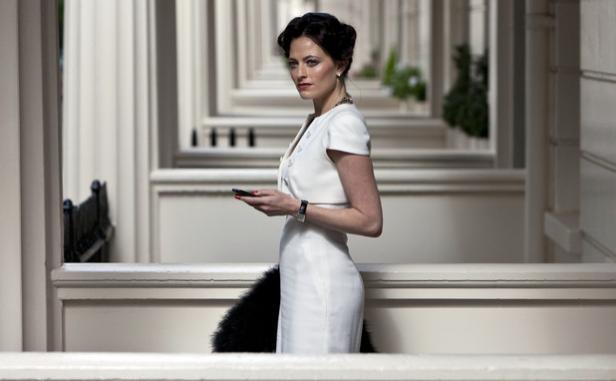 Eine Frau in einem weißen Kleid steht vor einer Reihe von Stadthäusern und blickt über ihre Schulter.