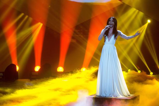 Conchita Wurst steht in einem weißen Kleid auf der Bühne und singt.