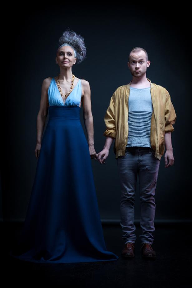 Eine ältere Frau in einem blauen Kleid hält Händchen mit einem jüngeren Mann in Jeans und Jacke.