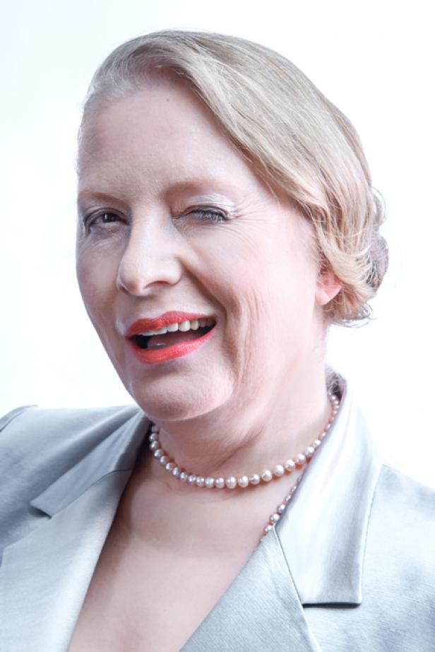 Eine Frau mit Perlenkette und Blazer zwinkert in die Kamera.