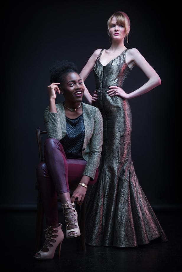 Zwei Frauen posieren in eleganter Kleidung vor einem dunklen Hintergrund.