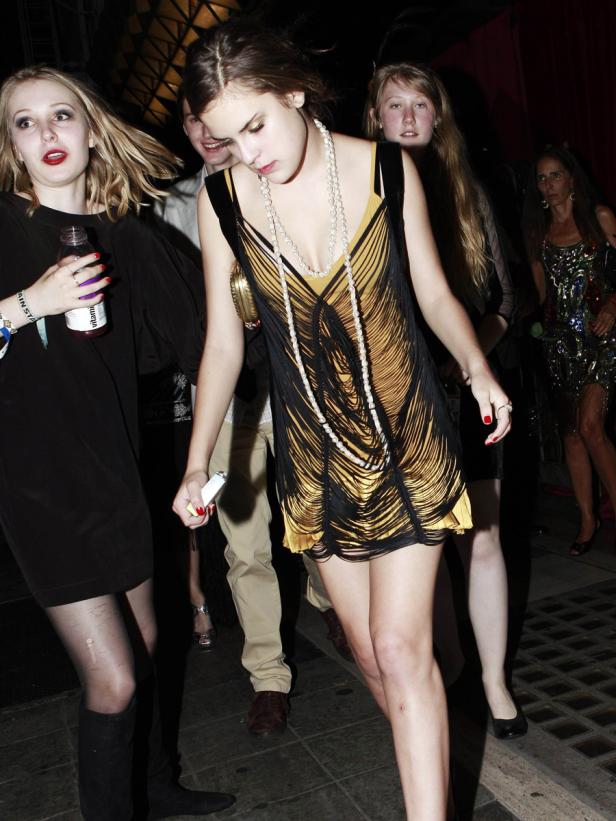 Eine junge Frau in einem gelb-schwarzen Fransenkleid und einer Perlenkette auf einer Party.