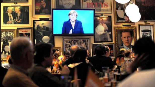 Menschen in einer Bar schauen Angela Merkel im Fernsehen zu.