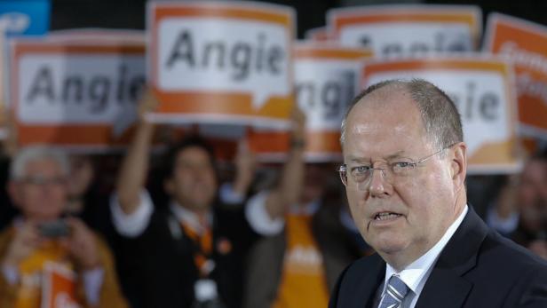 Ein Mann mit Brille spricht vor einer Menge, die Schilder mit der Aufschrift „Angie“ hochhält.