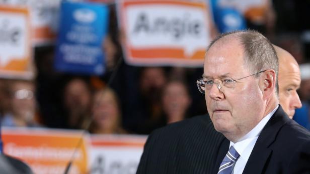 Ein Mann mit Brille und Anzug steht vor einer Menschenmenge mit Schildern.