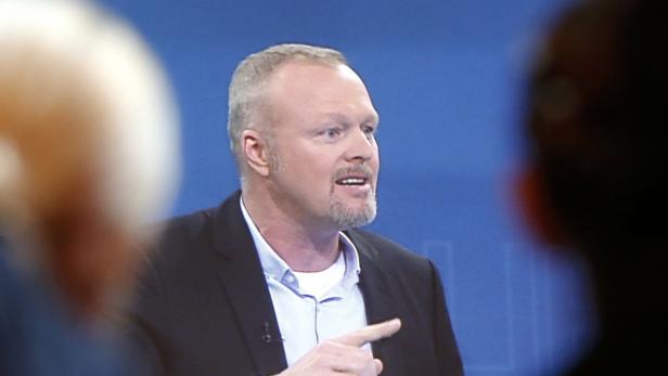 Ein Mann mit Bart gestikuliert vor einem blauen Hintergrund.