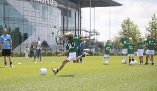 Ein Kind schießt auf einem Fußballfeld vor der Volkswagen Arena einen Ball.