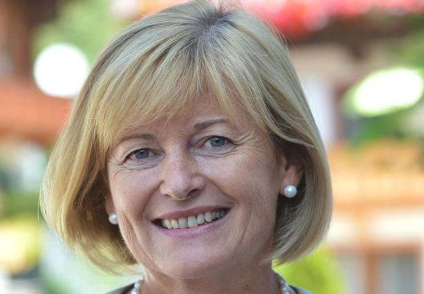 Porträt einer lächelnden Frau mit blonden Haaren und Perlenohrringen.