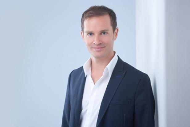 Ein Mann in einem blauen Sakko und einem weißen Hemd lehnt an einer Wand.