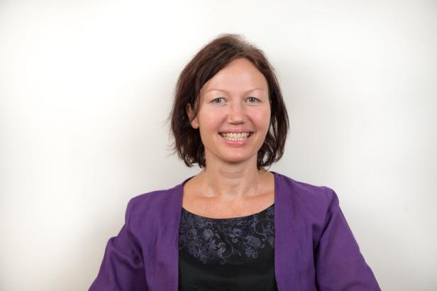 Eine lächelnde Frau mit braunen Haaren und einer lila Jacke vor einem weißen Hintergrund.