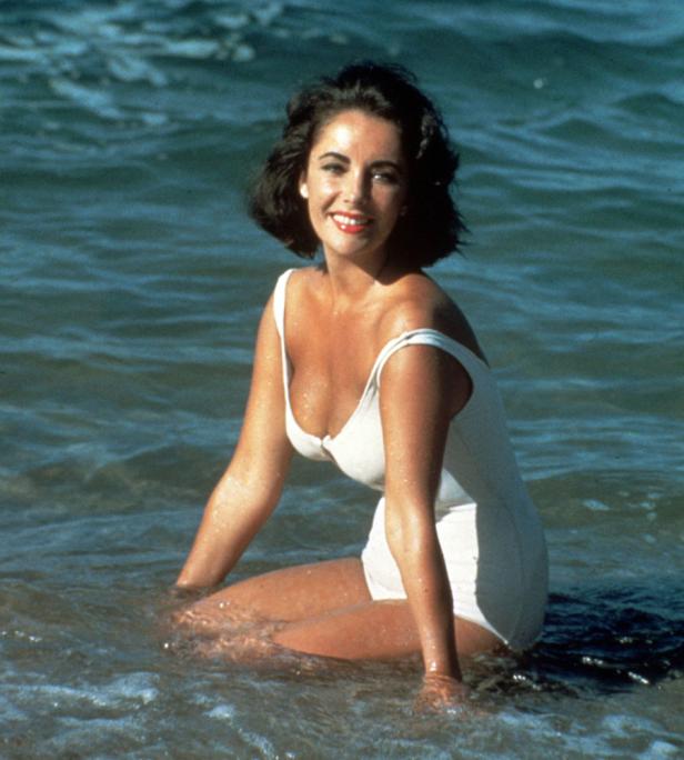 Elizabeth Taylor sitzt in einem weißen Badeanzug im Meer.