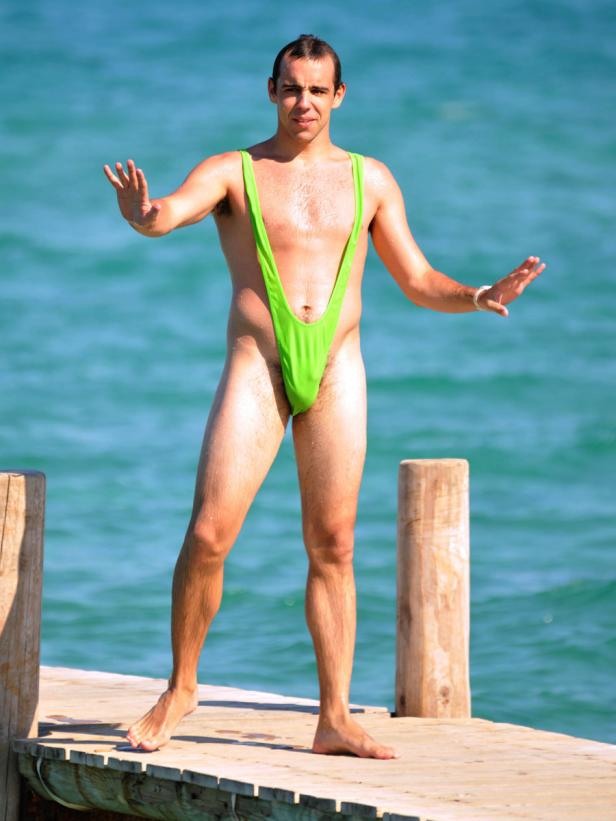 Ein Mann steht auf einem Holzsteg in einem grünen Mankini vor dem Meer.