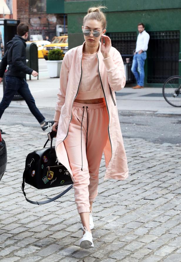 Gigi Hadid in einem rosa Trainingsanzug und mit einer schwarzen Reisetasche auf einer Kopfsteinpflasterstraße.