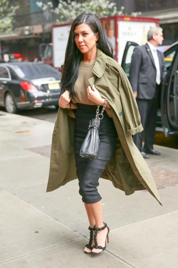 Kourtney Kardashian mit olivgrünem Trenchcoat, schwarzem Rock und Sandalen auf dem Bürgersteig.