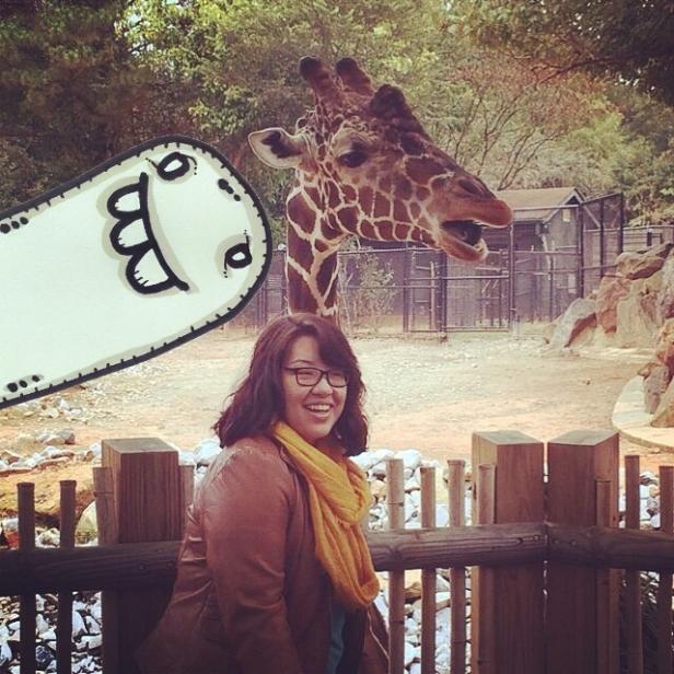 Eine Frau lächelt vor einem Zaun, mit einer Giraffe und einem Cartoon im Hintergrund.