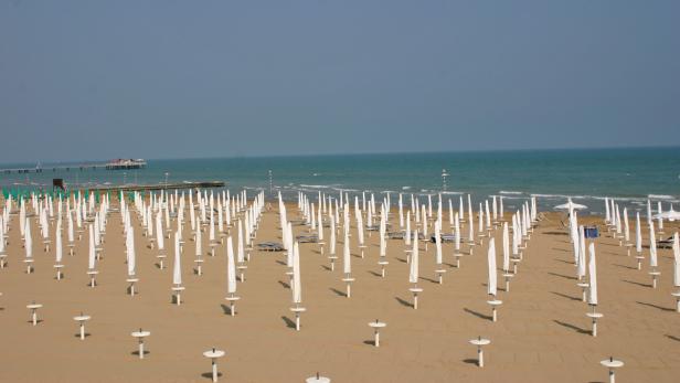 Ein Strand mit vielen geschlossenen, weißen Sonnenschirmen und einem Pier im Hintergrund.