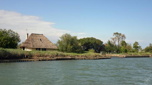 Ein Haus mit Strohdach am Ufer eines Gewässers.