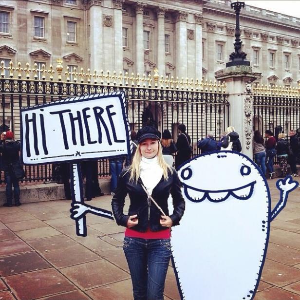Eine Frau posiert mit einem Schild mit der Aufschrift „Hi There“ und einer Cartoon-Figur vor dem Buckingham Palace.
