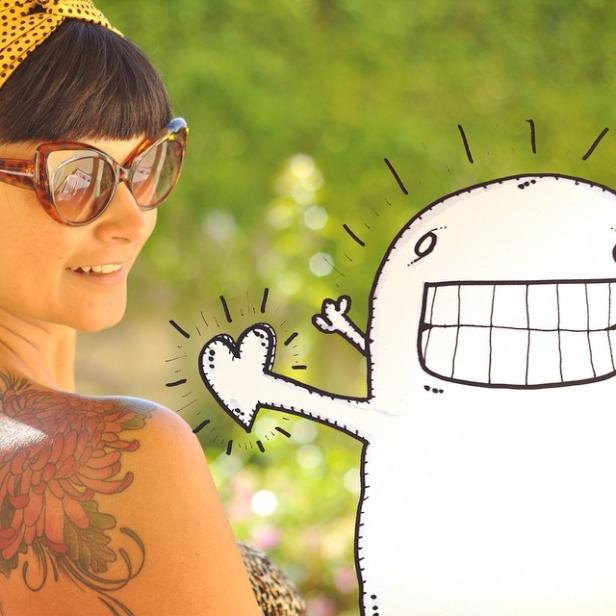 Eine Frau mit Sonnenbrille und Tattoo posiert mit einer gezeichneten Comicfigur.
