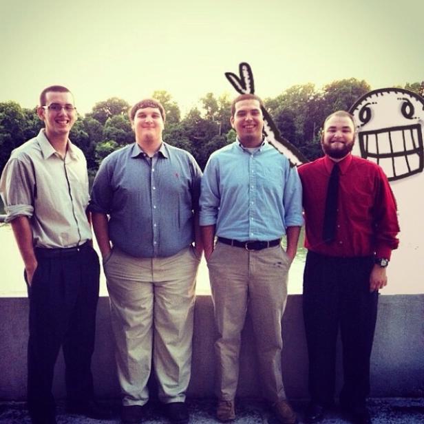 Vier Männer posieren vor einer gezeichneten Figur mit einem breiten Grinsen.