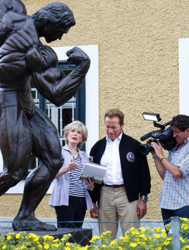 Arnold Schwarzenegger steht mit einer Frau und einem Kameramann vor einer Statue.