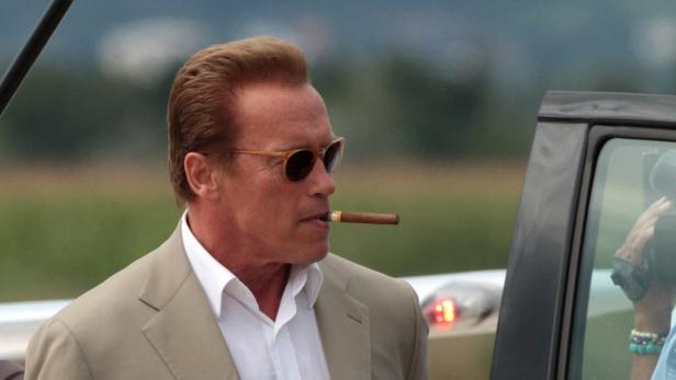 Arnold Schwarzenegger mit Zigarre im Mund, neben einem Auto stehend.