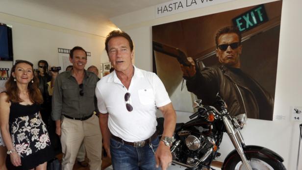 Arnold Schwarzenegger posiert vor einem „Terminator“-Poster und einem Motorrad.