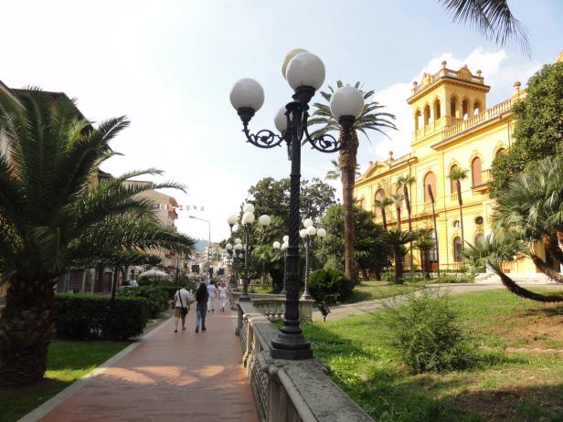 Ein gelbes Gebäude mit einem Turm und Palmen an einer Promenade.