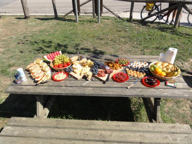 Ein Picknicktisch ist reichlich mit Speisen wie Wurst, Käse, Obst und Brot gedeckt.