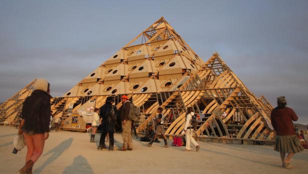 Besucher stehen vor einer großen, pyramidenförmigen Holzkonstruktion in der Wüste.