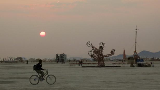 Eine Person fährt mit dem Fahrrad durch die Wüste bei Sonnenuntergang, im Hintergrund Kunstinstallationen.
