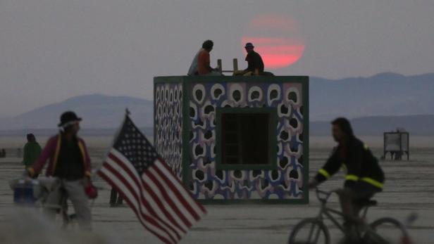 Auf einer staubigen Ebene stehen Menschen und eine amerikanische Flagge vor einem bemalten Gebäude im Abendlicht.