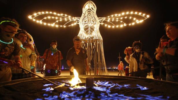 Menschen versammeln sich um ein Feuer vor einer beleuchteten Engelsstatue auf dem Burning Man Festival.