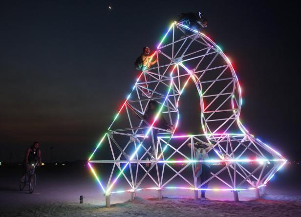 Menschen klettern auf einer leuchtenden, geometrischen Skulptur unter einem Nachthimmel.